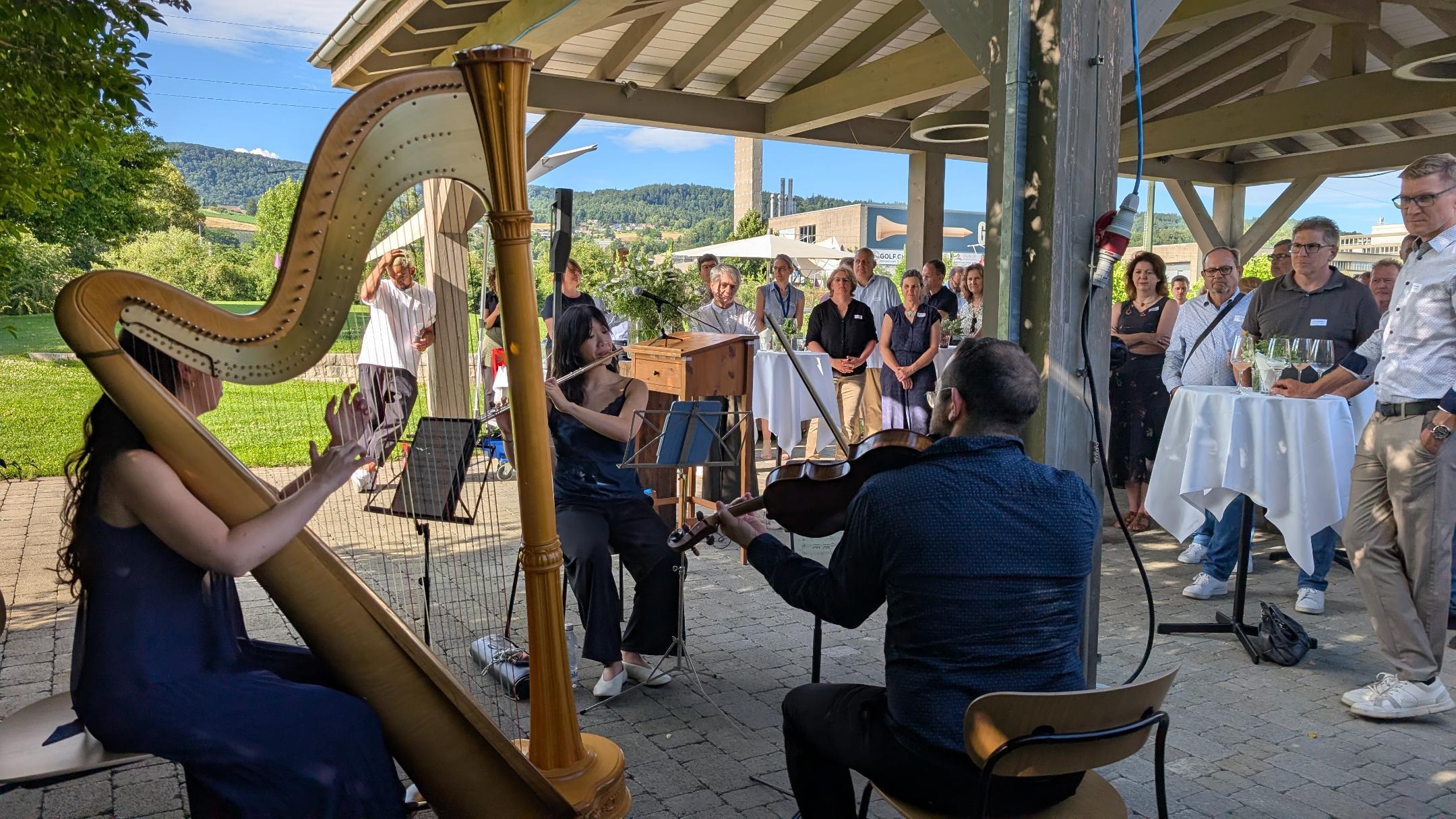 Ein Musiktrio mit Harfe, Querflöte und Geige spielt im Freien vor einer Gruppe von Gästen.