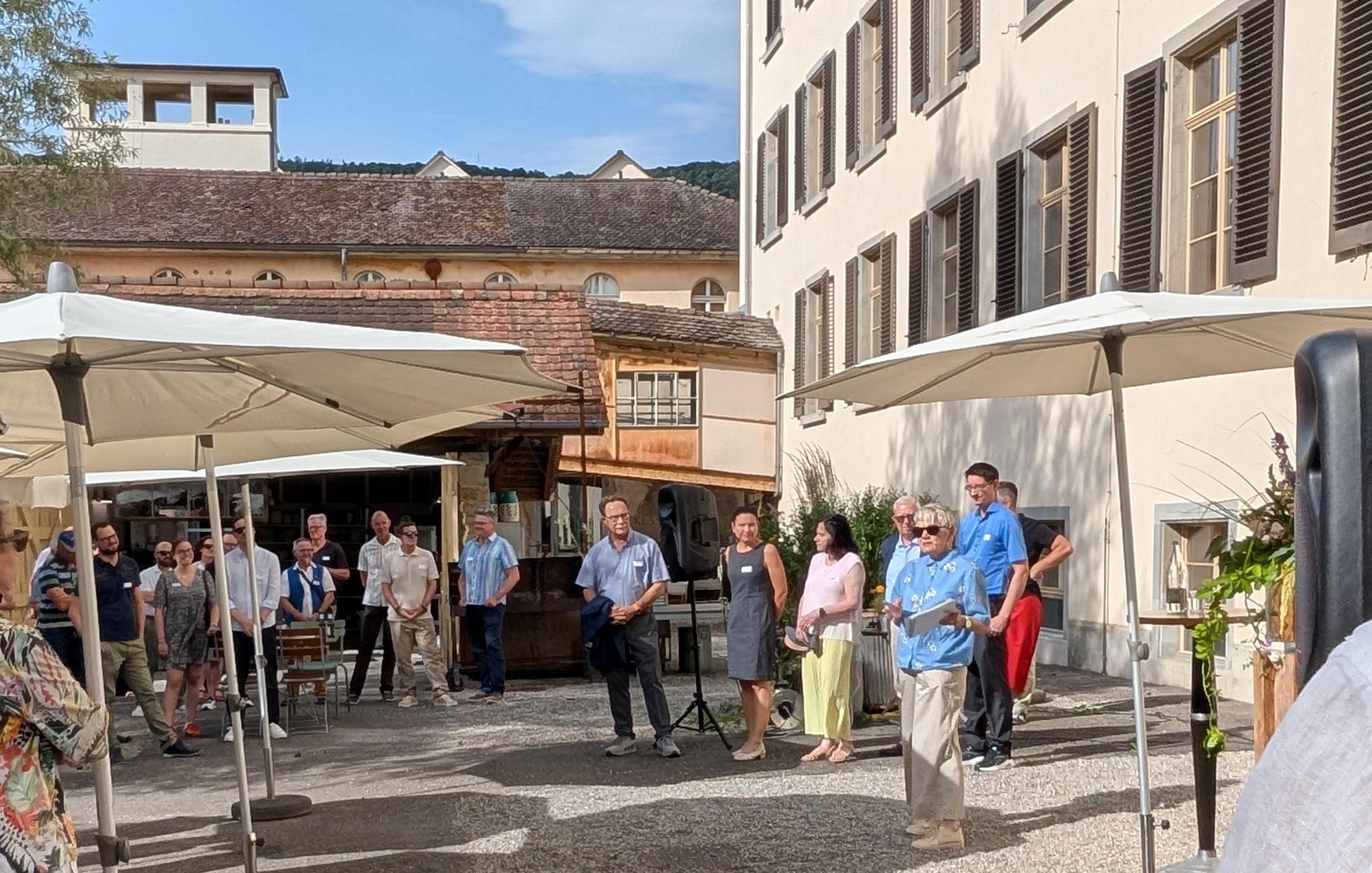 Blick auf einen Innenhof, in dem links Zuhörende stehen und rechts eine Gruppe von sechs Menschen zu ihnen positioniert ist.