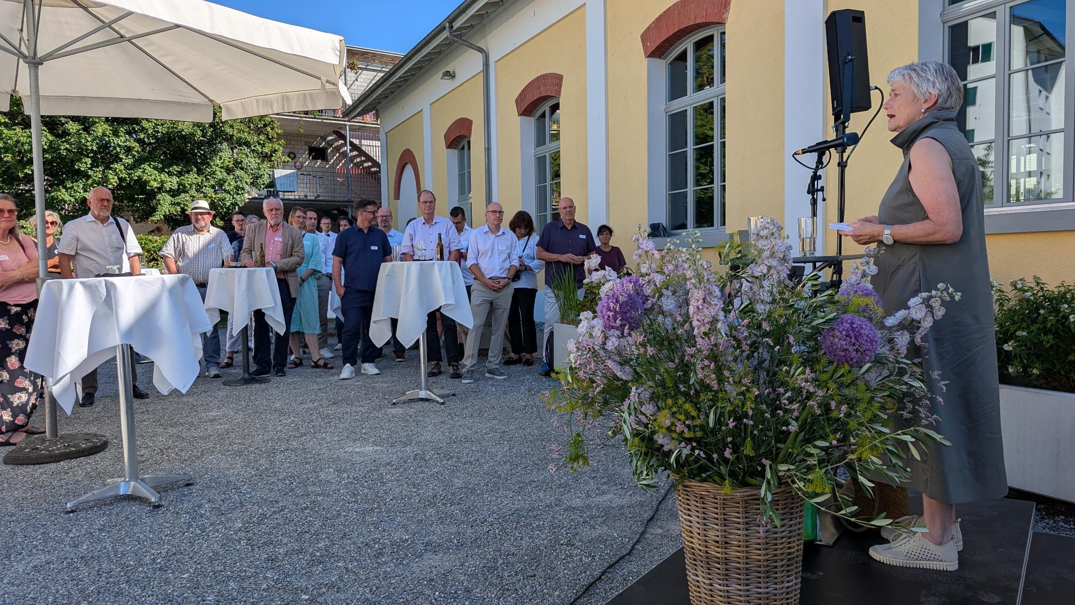 Rechts eine Frau auf einem Podium, die zu einer Gruppe von rund 50 Menschen spricht.