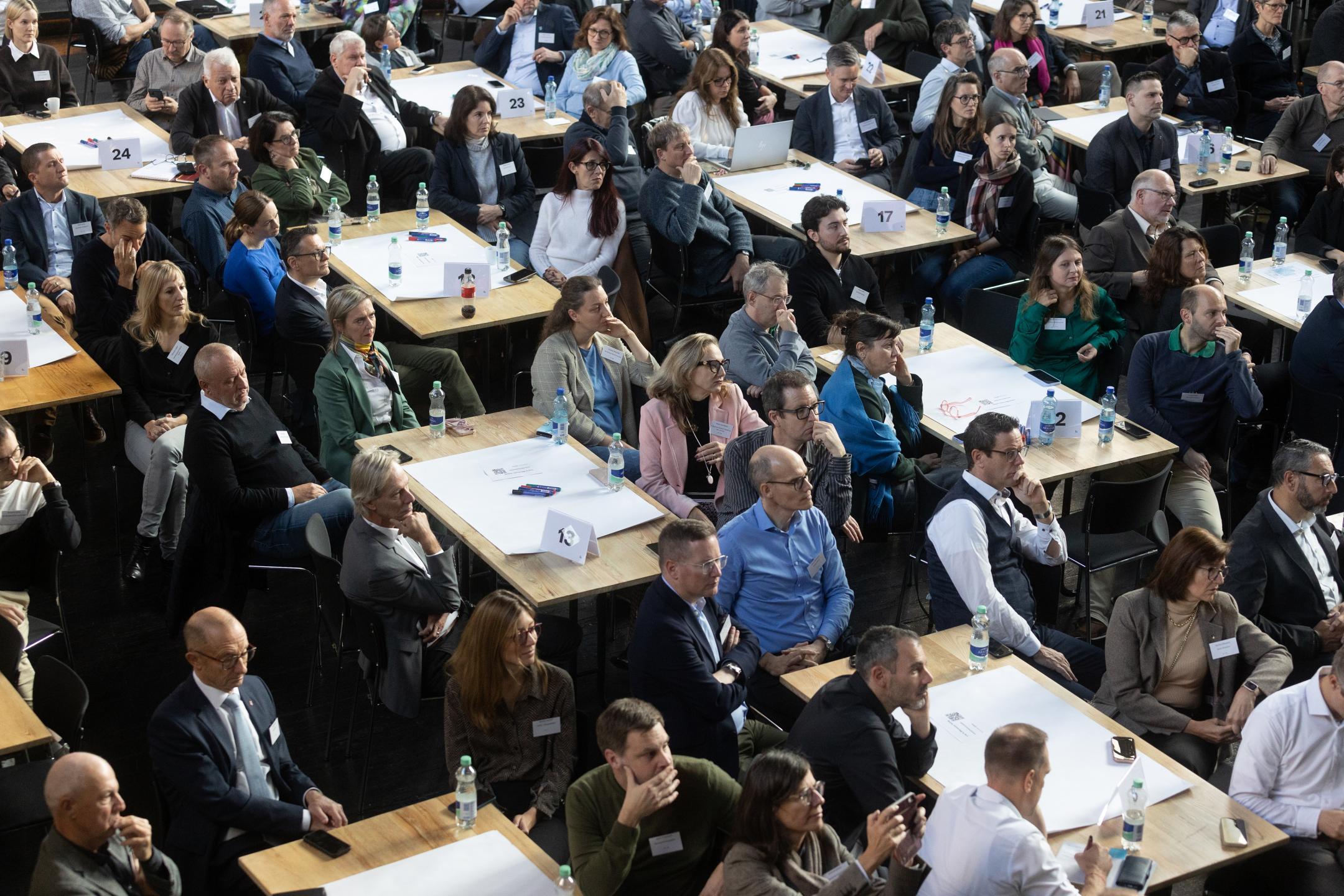 Ein Saal, in dem viele Menschen um Tische herum sitzen und in eine Richtung schauen.