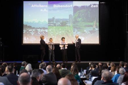 Vier Personen stehen auf der Bühne und diskutieren miteinander, im Hintergrund eine Leinwand mit je einem Landschaftsbild von Affoltern a.A., Dübendorf und Zell.