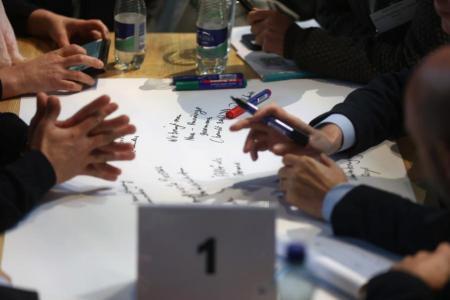 Nahaufnahme von gestikulierenden Händen und einer Tischfläche, auf der Papier und Stifte liegen. 