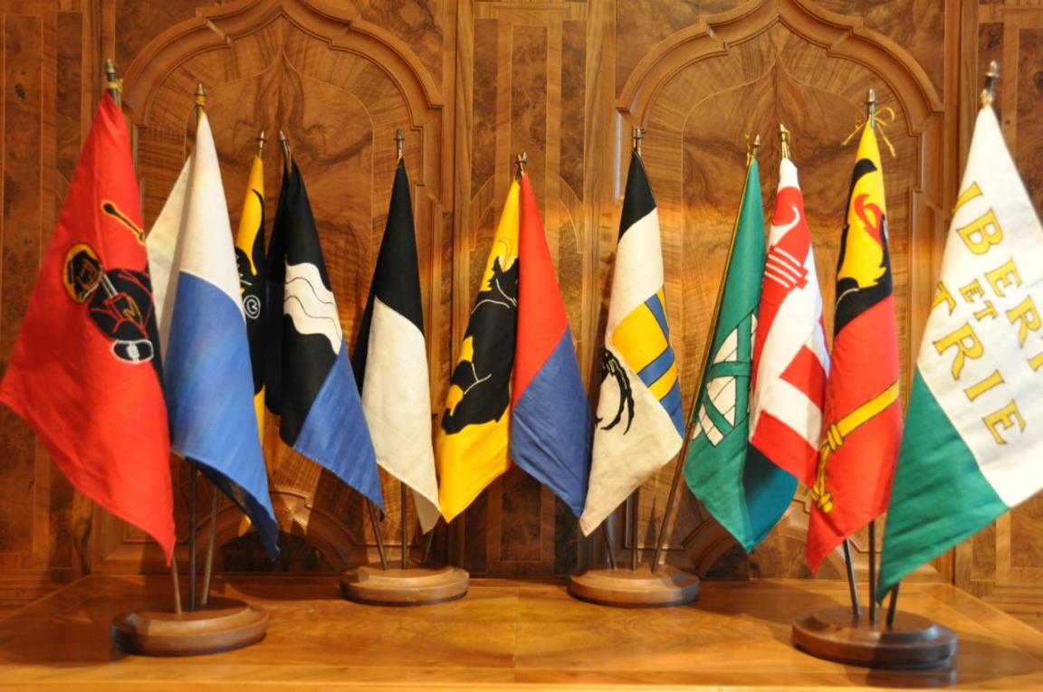 Kleine Fahnen der Kantone Glarus, Luzern, Uri, Aargau, Fribourg, Bern, Tessin, Graubünden, St.. Gallen, Jura, Genf und Waadt auf einem Holztisch stehend als Symbol für die interkantonale Zusammenarbeit