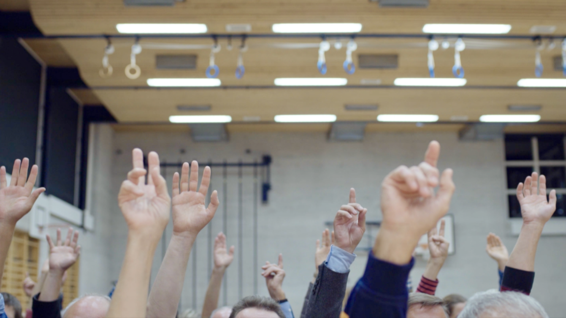 Bei einer Gemeindeversammlung in einer Turnhalle strecken Stimmberechtigte Ihre Hände in die Luft, um einer Vorlage zuzustimmen. 