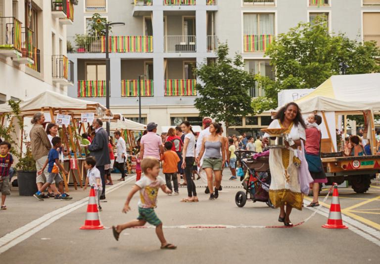 Das Bild zeigt in lebendiges Strassenfest in einem Wohnquartier mit Marktständen, spielenden Kindern und fröhlichen Menschen. Eine Frau trägt lächelnd eine Schale mit Essen, während andere Besucherinnen und Besucher flanieren oder sich unterhalten. Die Balkone und Fenster der Häuser im Hintergrund sind mit bunten Stoffen geschmückt.