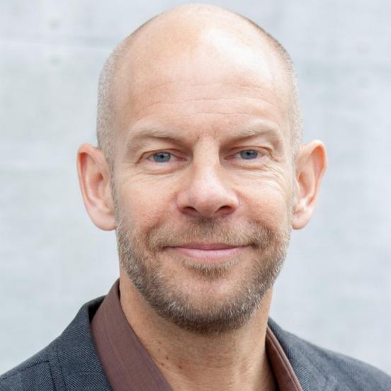 Ein Mann mit kurzen Haaren, braunem Hemd und grauem Blazer