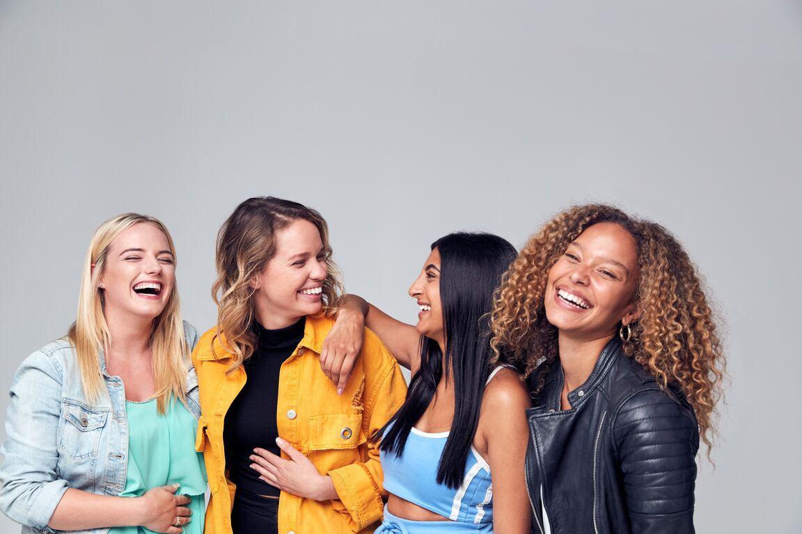 Vier junge Frauen stehen vor einem grauen Hintergrund und lachen.