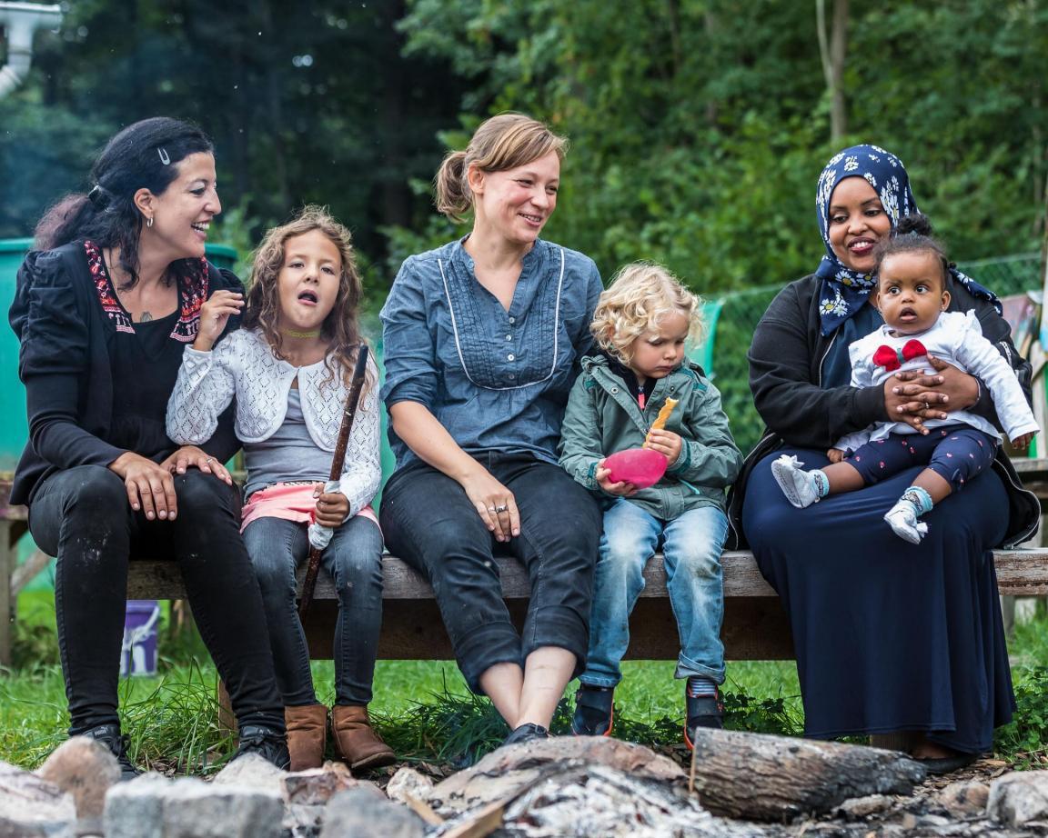 Drei Frauen sitzen mit ihren Töchtern nebeneinander auf einer Bank vor einem Wald.
