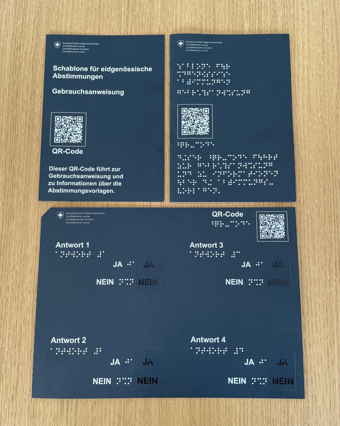 Auf einem braunen Holztisch sind die Abstimmungsschablone und je eine Gebrauchs-anweisung in Druckschrift und eine Brailleschrift aufgelegt. Die Abstimmungsschablone und die Gebrauchsanweisungen sind aus schwarzem Karton gefertigt. Bedruckt sind sie mit weisser grosser Druckschrift und mit Brailleschrift. Zudem ist sowohl auf der Schablone als auch auf den Gebrauchsanweisungen ein QR-Code aufgedruckt.