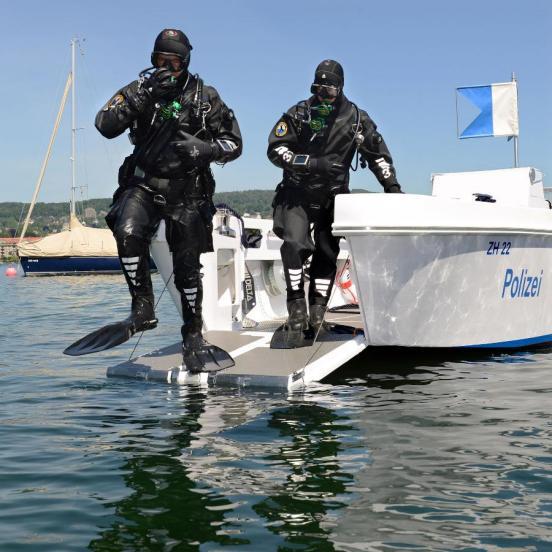 Zwei Personen in Tauchanzügen und Schwimmflossen springen von einer heruntergelassenen Heckklappe eines Bootes der Stadtpolizei in den Zürichsee.