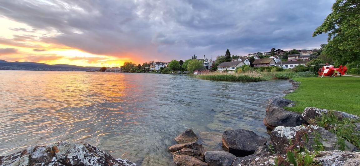 Das Foto zeigt Abendstimmung mit Sonnenuntergang am Zürichsee. Auf einer Wiese am Wasser steht ein Helikopter der Schweizerischen Rettungsflugwacht.