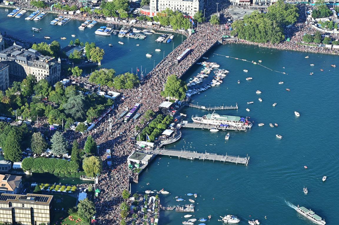 Drohnenaufnahme zeigt die Zürcher Bellevuebrücke von oben. Die Brücke ist voller Menschen, erkennbar an einer Vielzahl heller Punkte. Im Seebecken rund um die Brücke sind sehr viele Boote. auf dem Wasser.