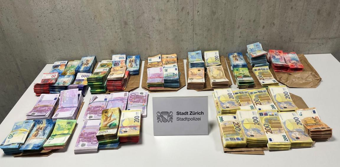 Sichergestellte Bargeldbeträge in verschiedenen Währungen liegen gebündelt auf einem Tisch. In der Mitte steht ein Schild der Stadtpolizei Zürich.