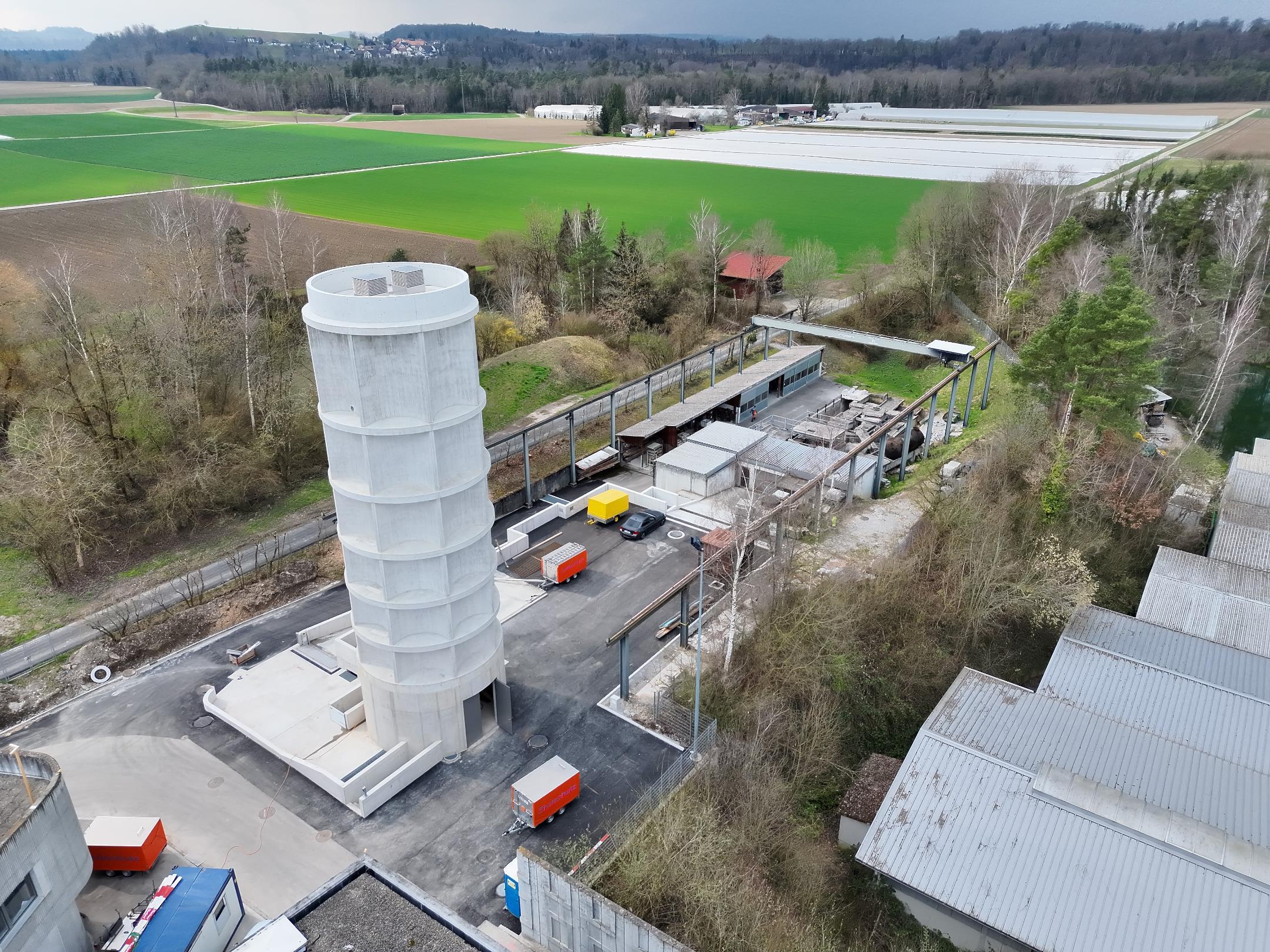 Es handelt sich um eine Luftaufnahme der Flutungsanlage - ein rund 23 Meter hoher Turm. Rundherum stehen mehrere Anhänger des Zivilschutzes. Dahinter sieht man mehrere grüne Felder und Wiesen.