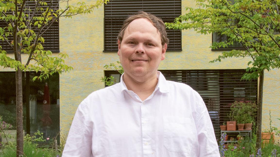 Kevin Rauch ist ein junger Mann mit kurzen, braunen Haaren, der vor einem Wohnhaus steht. Er trägt ein weisses Hemd.