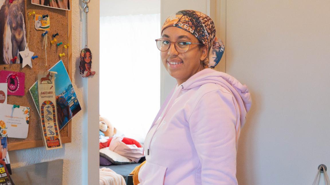 Das Bild zeigt eine junge Frau im Gang ihrer Wohnung. Sie trägt eine Brille, ein Kopftuch und einen rosafarbenen Kaputzenpulli.