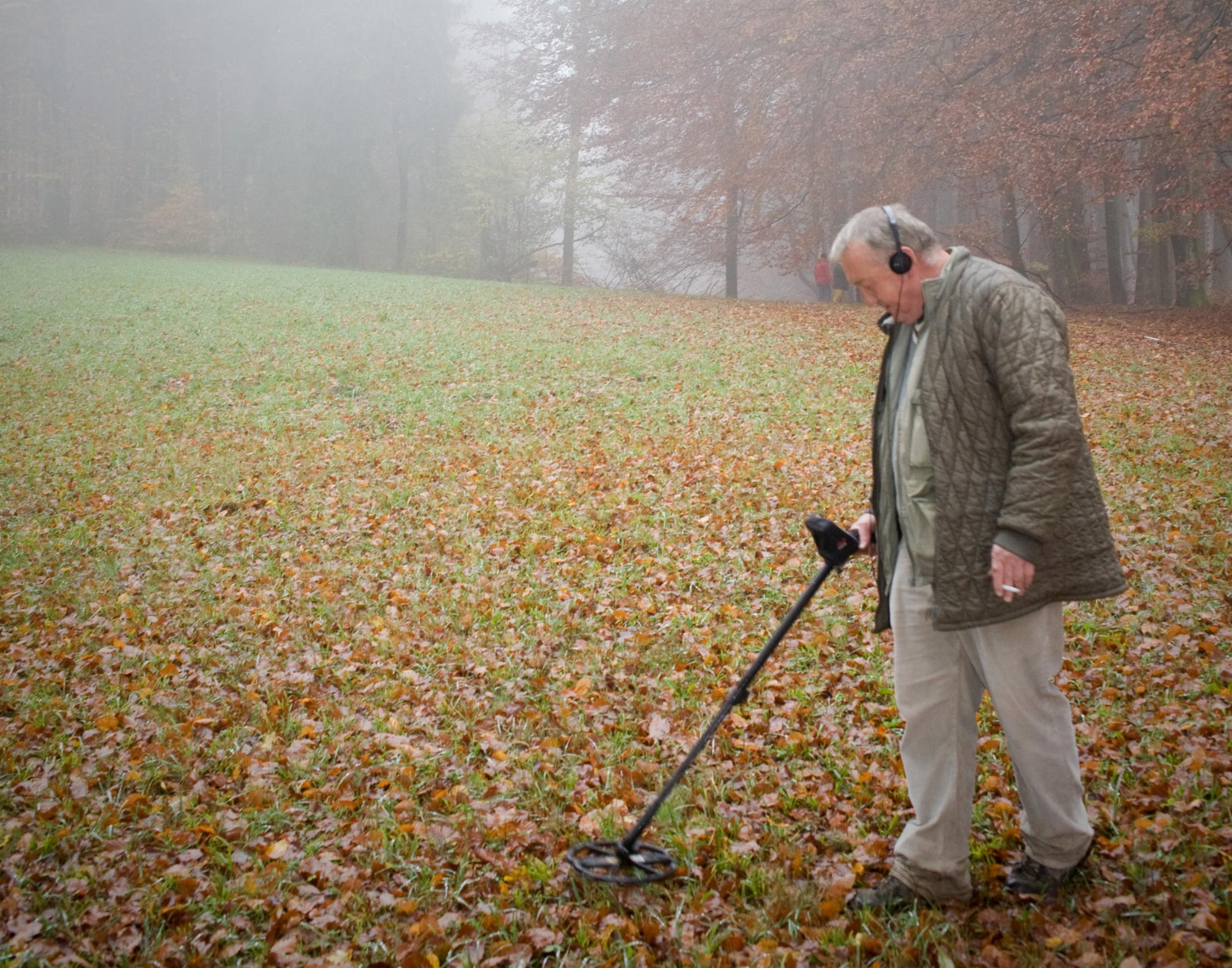 Ein Mann sucht mit einem Metaldetektor den Boden einer Lichtung ab.