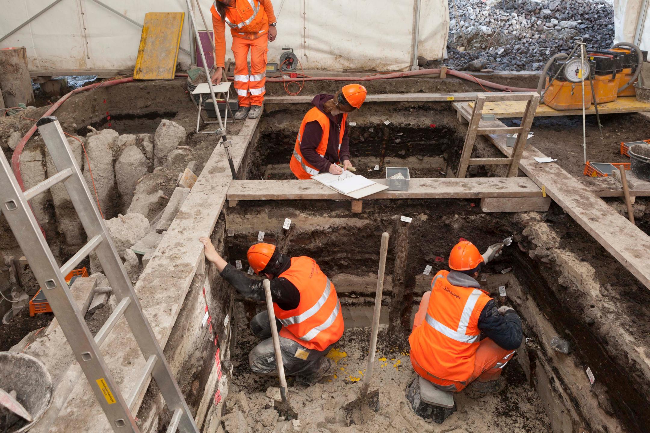 Ausgräber und Ausgräberinnen graben in einem Zelt. Eine Frau erfasst die Fundstelle mit einem Stift.