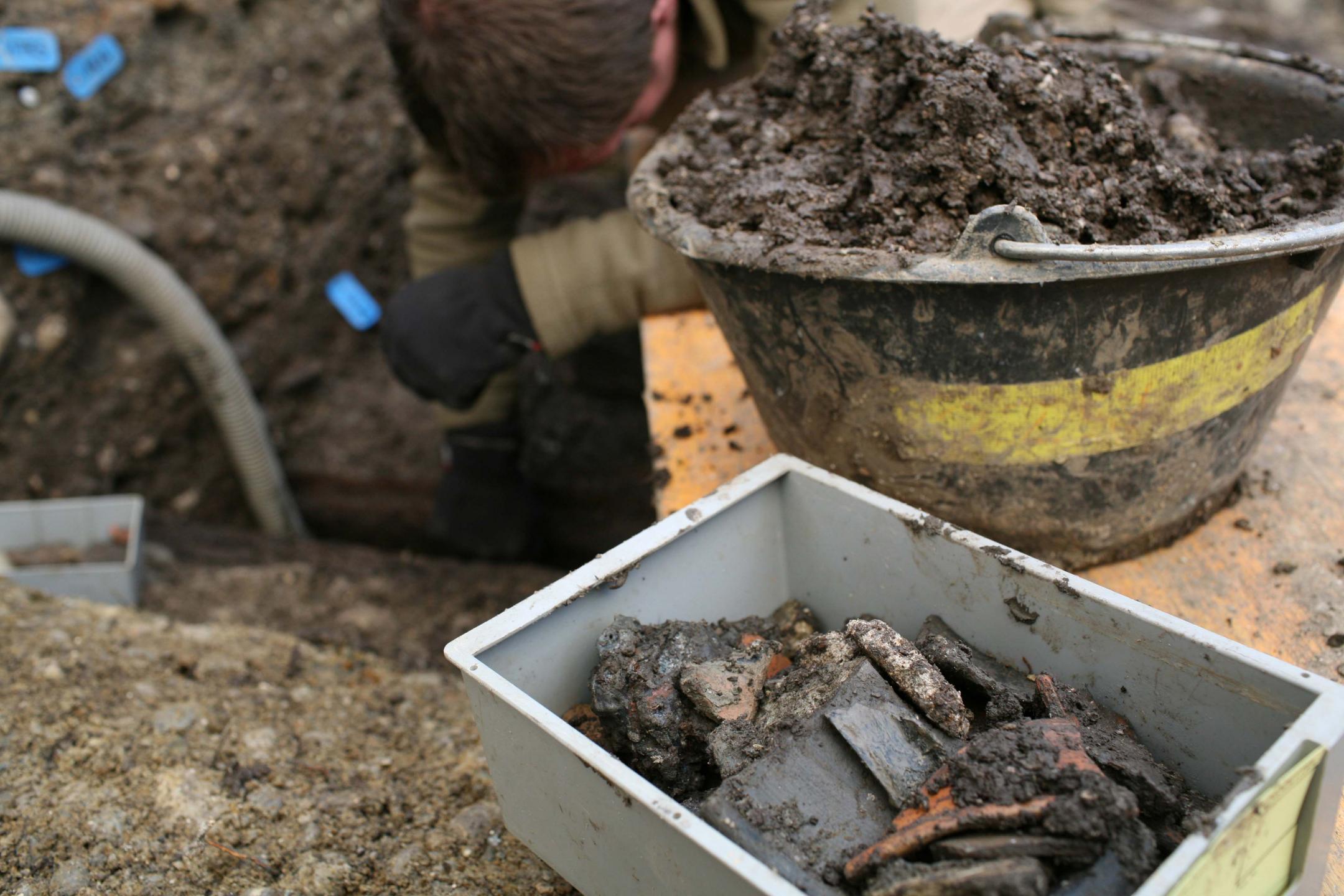 Im Vordergund sind gefundene Scherben in einer Box abgelegt. Im Hintergrund gräbt ein Mann.
