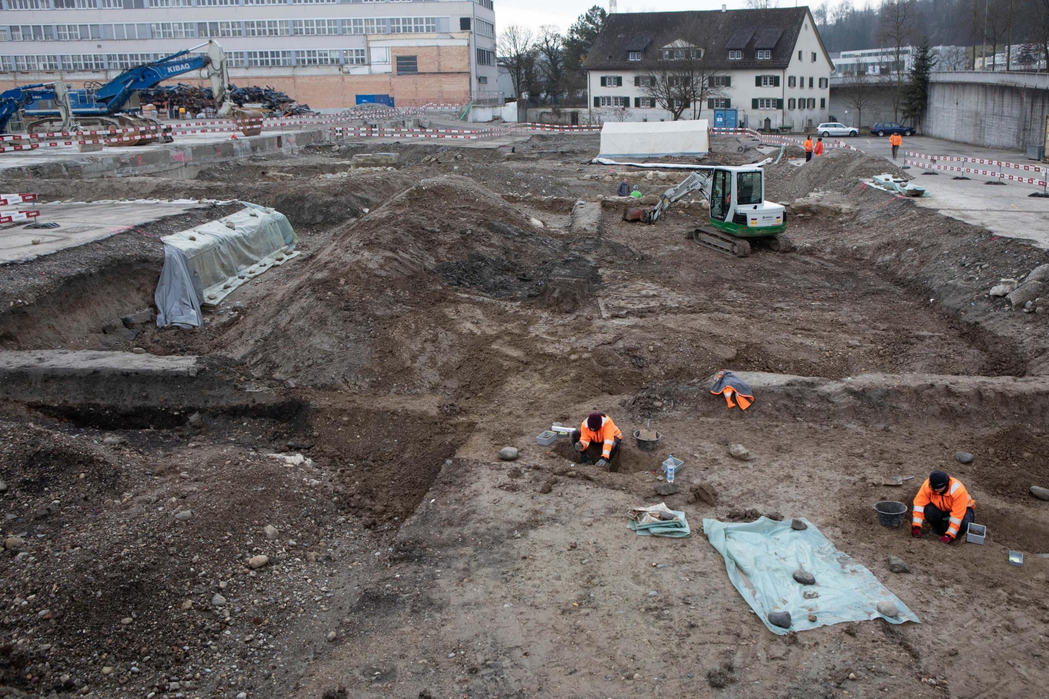 Eine archäologische Grabung mit zwei Personen im Vordergrund, die graben. Im Hintergrund sind Bagger und ein Industriegelände zu erkennen.