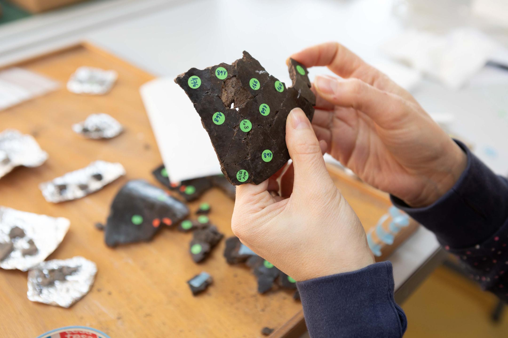 Scherben werden von zwei Hände zu einem grösseren Bruchstück eines Gefässes zusammengefügt. Farbige Klebepunkte helfen bei der Orientierung.
