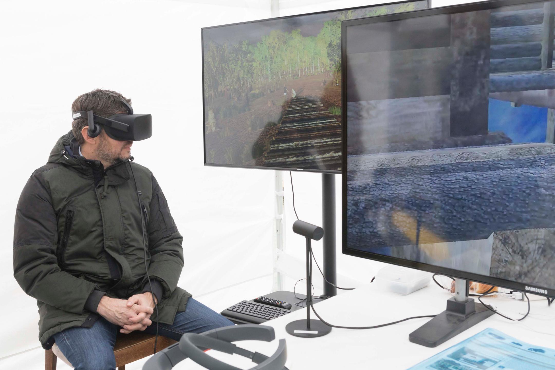 Ein Mann trägt eine VR-Brille und sitzt auf einem Stuhl. Daneben ist auf zwei Bildschirmen die Rekonstruktion einer Pfahlbauersiedlung zu sehen. 