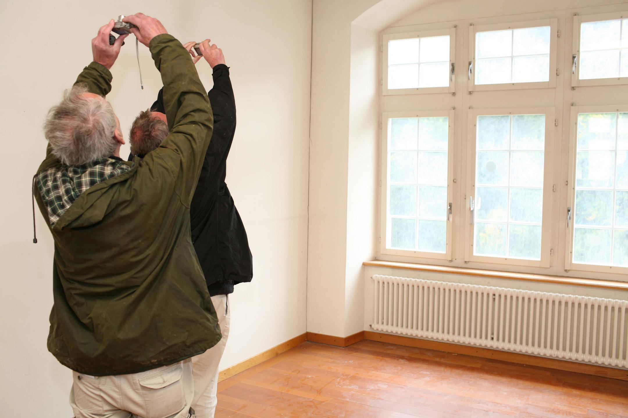 Zwei Männer stehen hintereinander in einem weissen Raum und fotografieren mit je einem Fotoapparat die Decke. Ihnen gegenüber liegt eine Fensterfront.