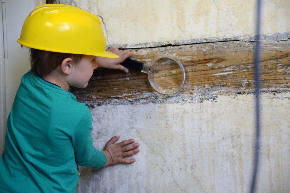 Ein Junge mit Bauhelm untersucht mit einer Lupe einen Holzbalken.