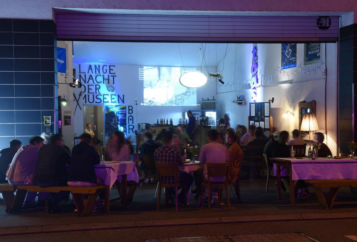 Gäste sitzen an Tischen in einer Garagenbar beim Alterthümer-Magazin