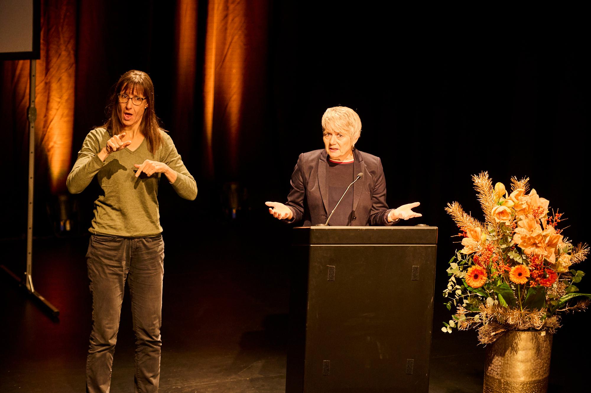 Jacqueline Fehr steht hinter Rednerpult, Geste mit offenen Armen, daneben die Übersetzerin, die in die Gebärdensprache übersetzt.