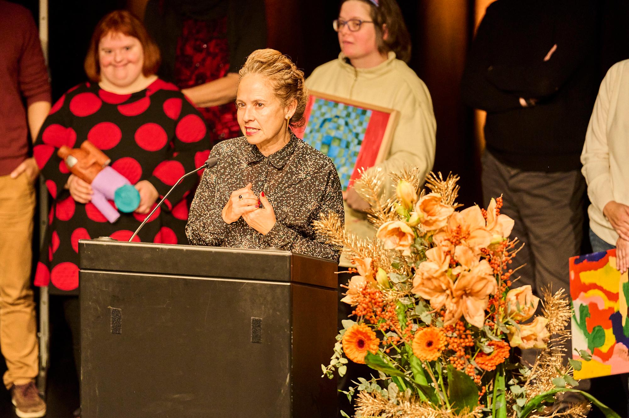 Kathrin Falk steht am Rednerpult und hält die Rede für die Kunstfabrik Wetzikon. Im Hintergrund steht die Gruppe der Kunstfabrik.