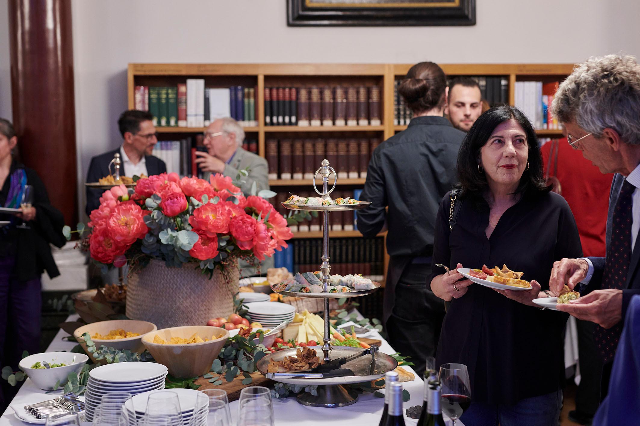 Ein Blick aufs Buffet mit feinen Köstlichkeiten und einem grosssen Strauss Pfingstrosen. Die Leute stehen darum herum und plaudern.