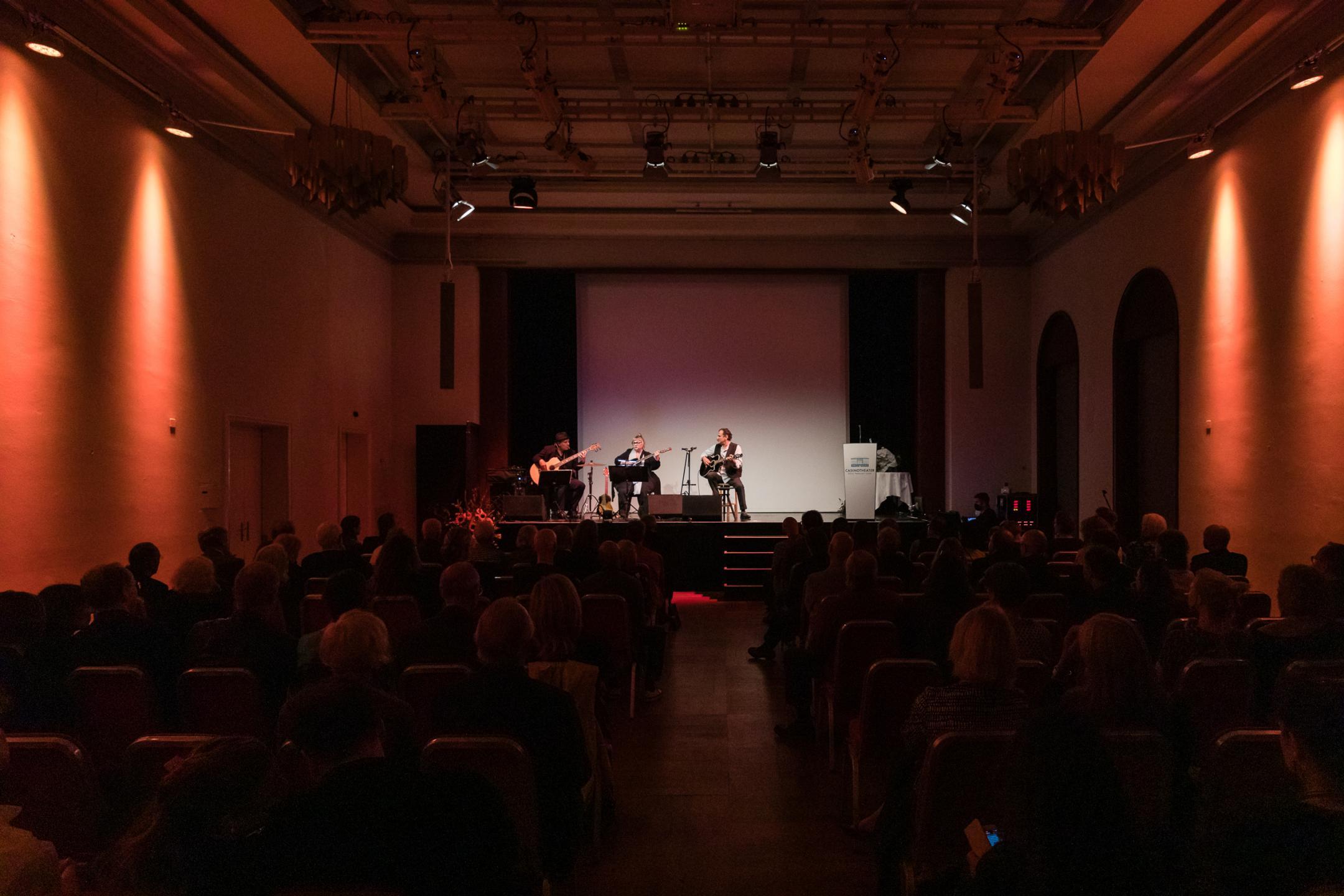 Blick von hinten im Saal Richtung Bühne. Dodo Hug und zwei Musiker spielen sitzend