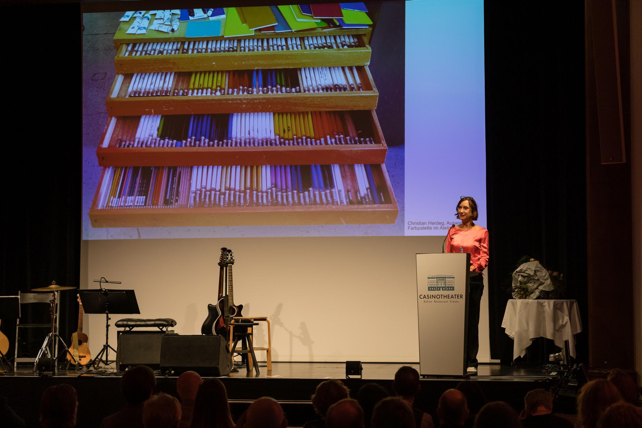 Sabine Schaschl am Rednerpult, im Hintergrund auf der Leinwand ein Schubladenmöbel mit vielen Siften und Pinseln