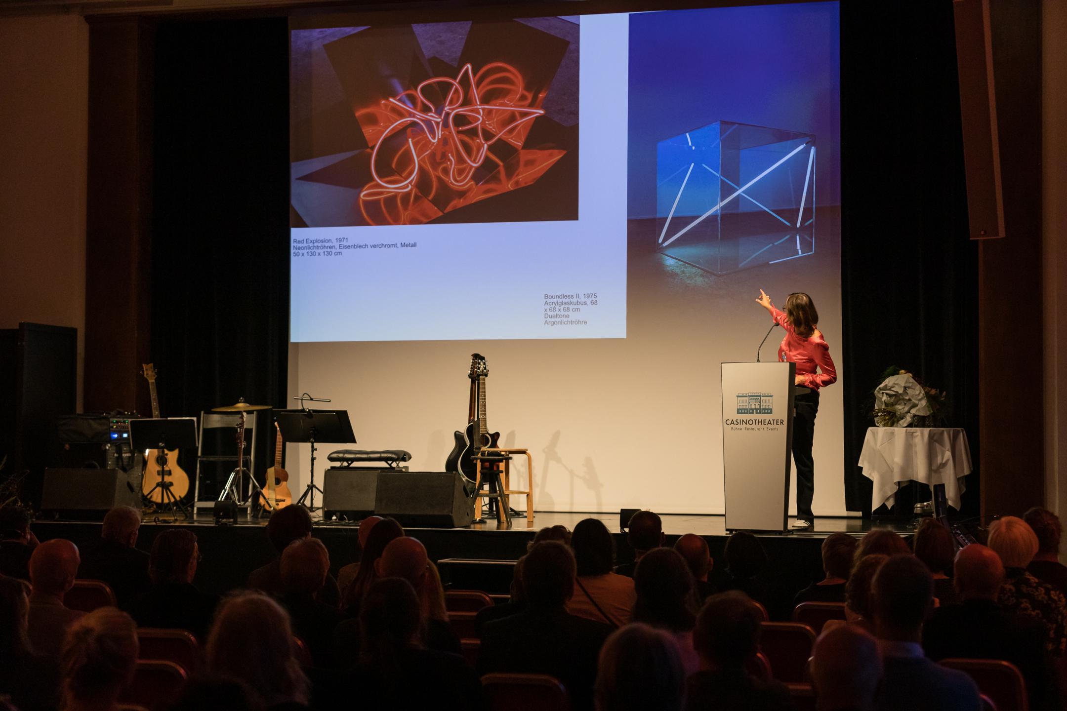 Sabine Schaschl am Rednerpult, im Hintergrund auf der Leinwand Installationen von Christian Herdeg