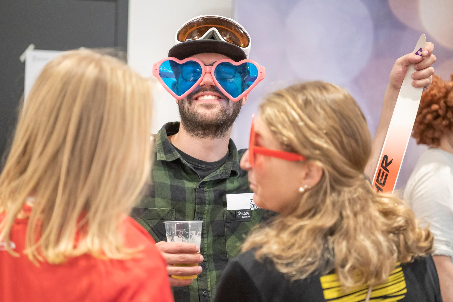 Ein Mann trägt eine grosse rosa Brille mit blauen Gläsern in Herzform. Drei Personen haben sich für ein Foto verkleidet. In einem Arm hält er zudem einen Langlaufski. 