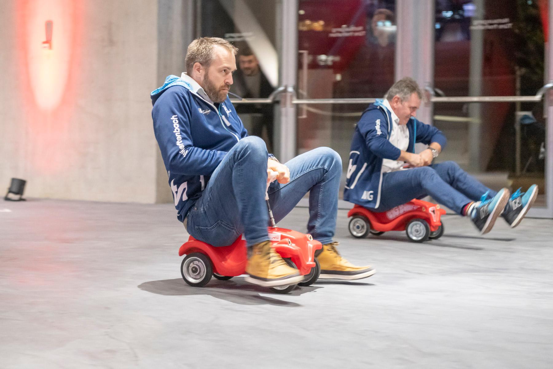 Zwei Herren fahren jeweils auf einem roten Bobbycar.