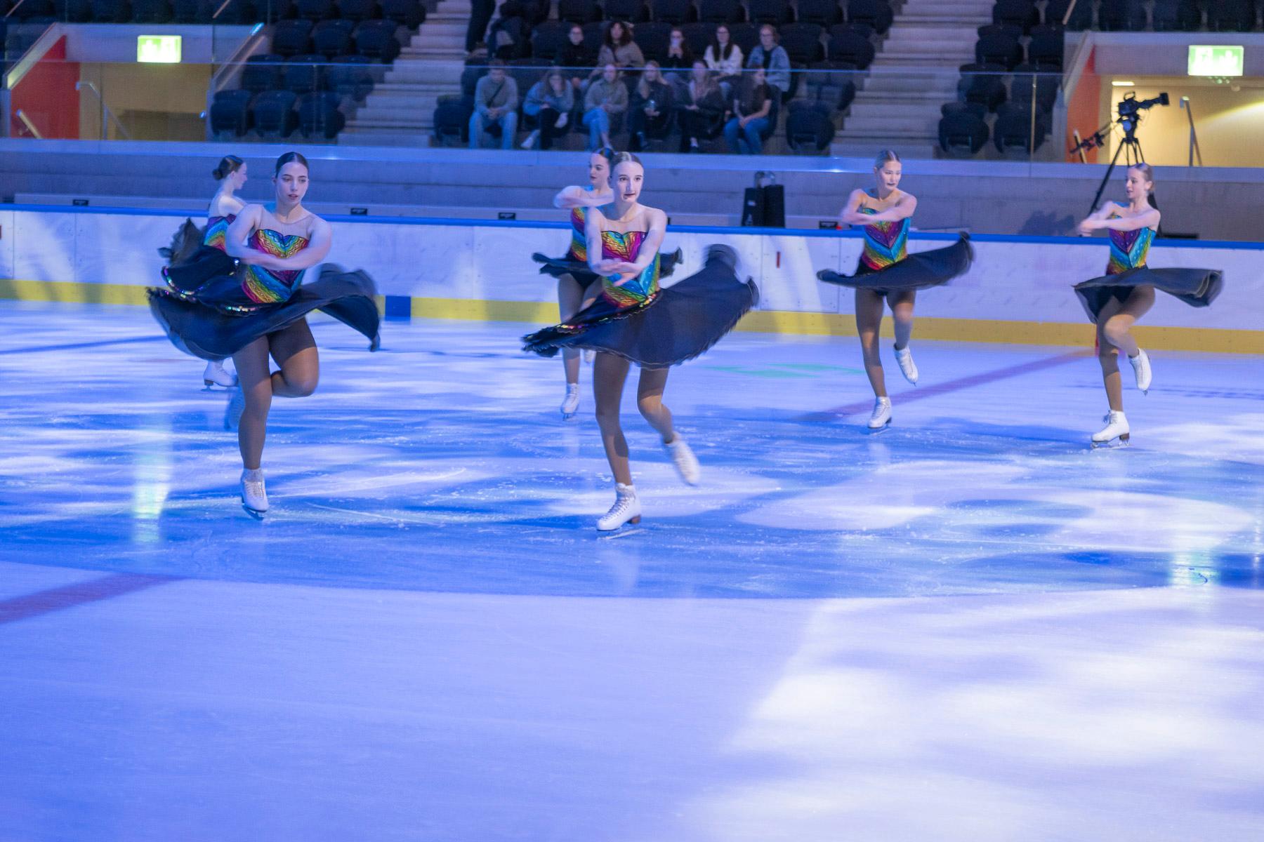Sechs Eiskunstläuferinnen mit regenbogenfarbigen Dress laufen synchron in einer Vorführung.