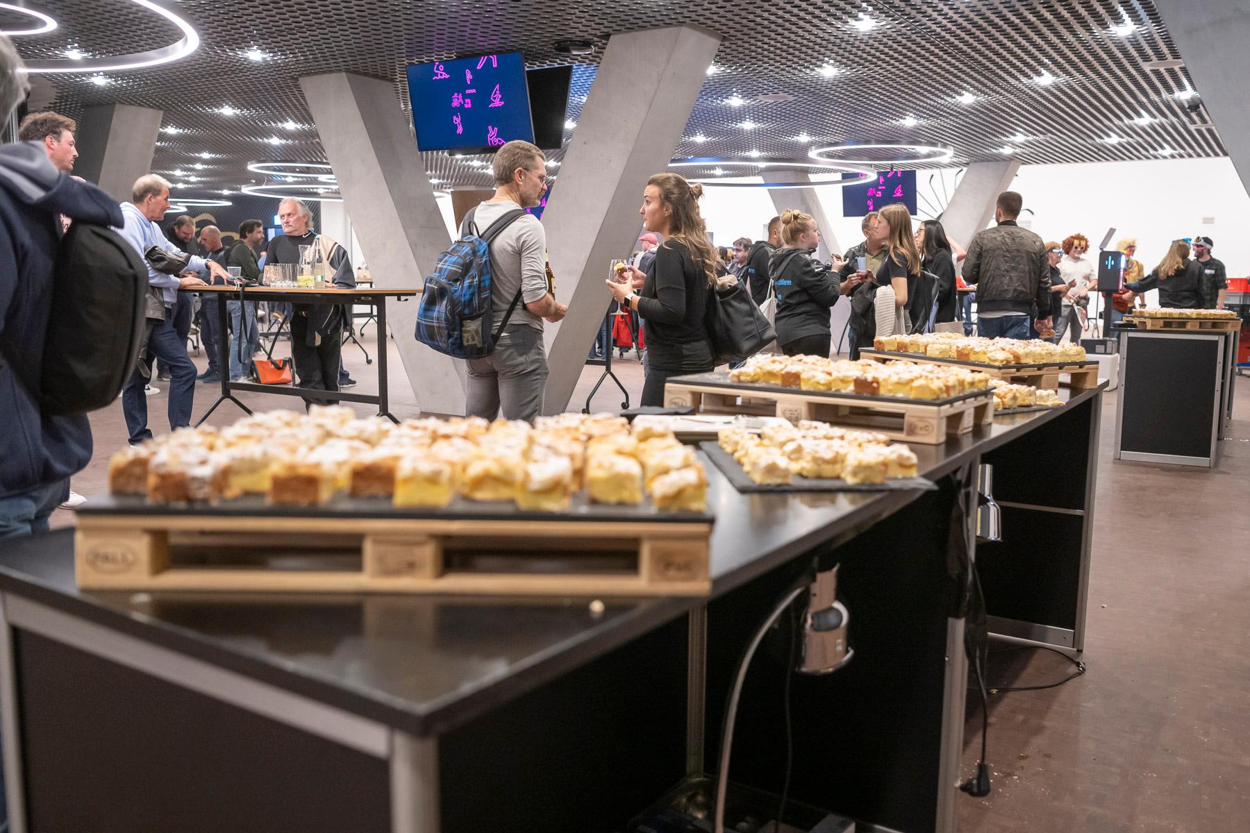 Im Business Club wurde ein Buffet mit Blechkuchen aufgebaut. Vor dem Buffet sind viele Menschen zu sehen, die in kleinen Gruppen zusammenstehen und miteinander sprechen. 