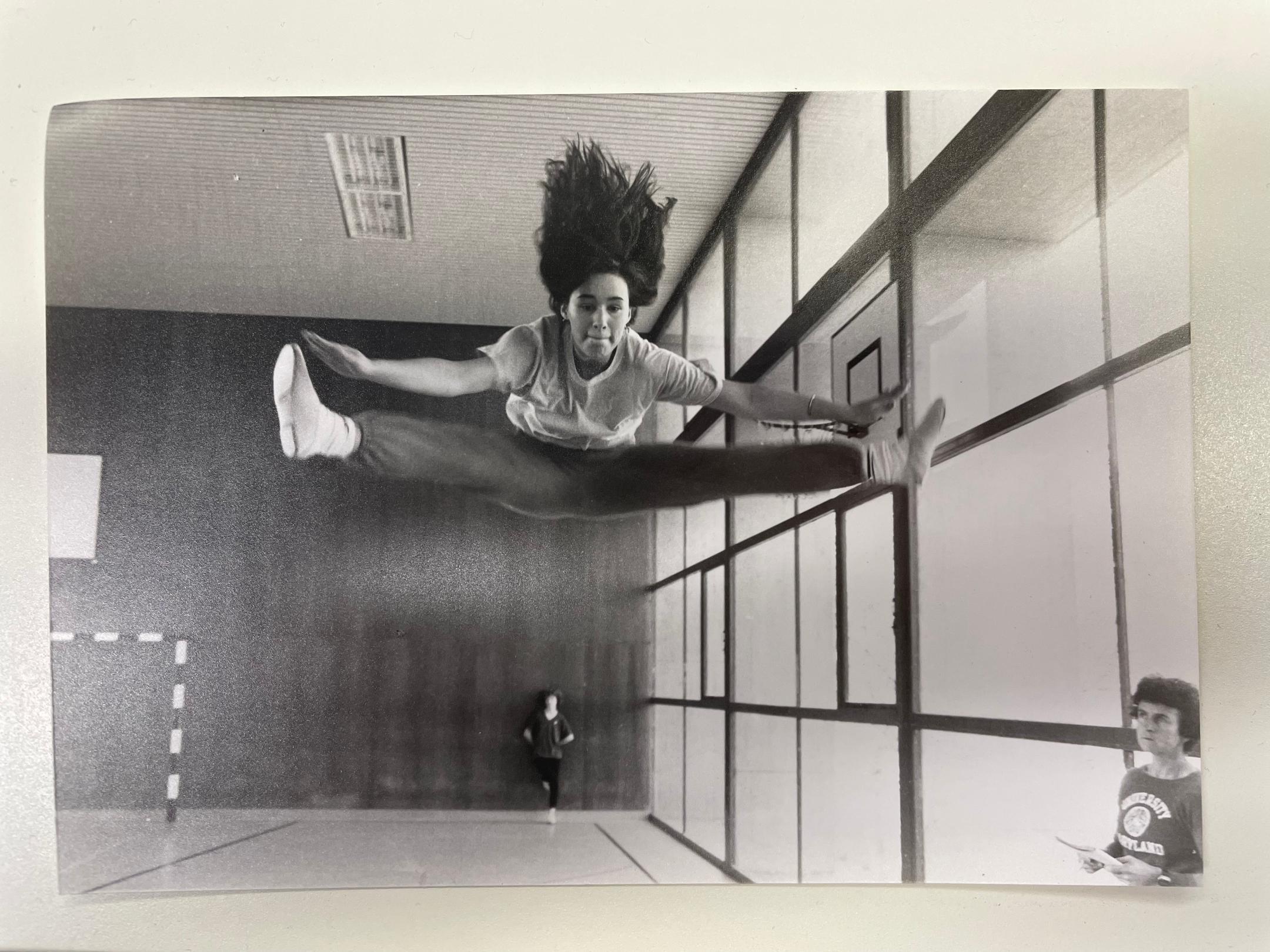 Schwarz-Weiss-Foto, dass in einer Turnhalle gemacht wurde und eine Jugendliche zeigt, die einen Grätschsprung macht.