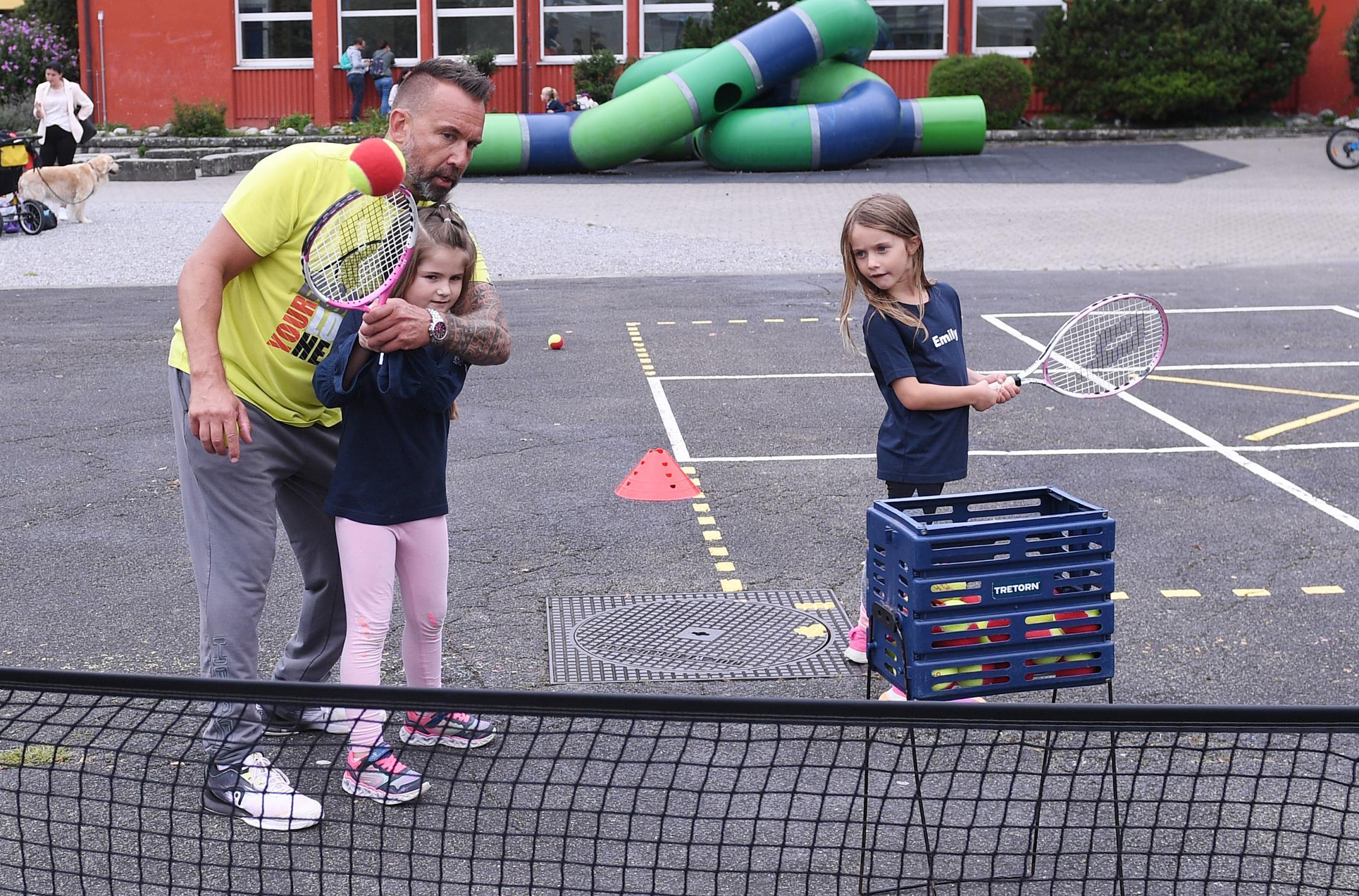 zwei Mädchen spielen auf dem Teerplatz Tennis und werden dabei von einem Trainer angeleitet