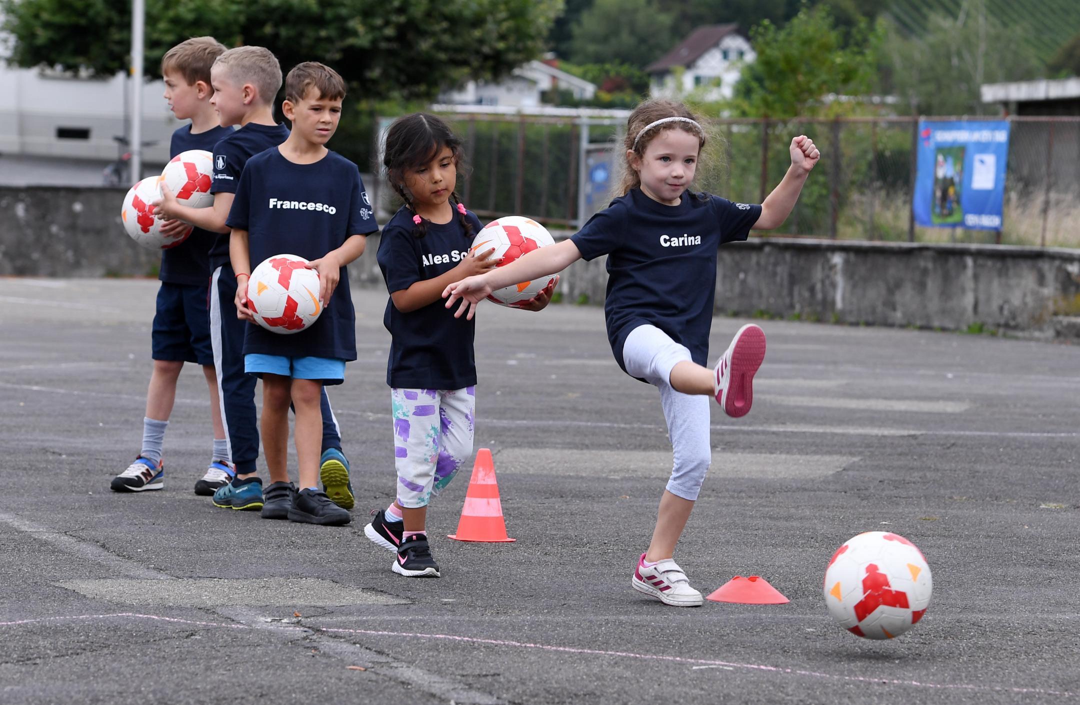 Ein Mädchen gibt auf dem Teerplatz einen Pass mit dem Fussball, Mädchen und Knaben warten in der Kolonne