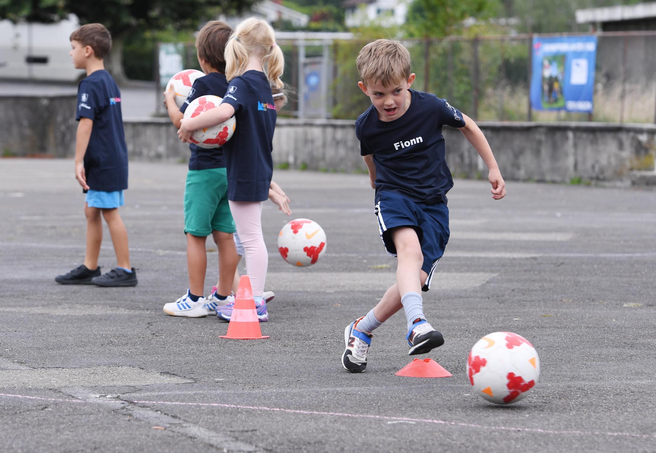 Ein Knabe gibt auf dem Teerplatz einen Pass mit dem Fussball, Mädchen und Knaben warten in der Kolonne
