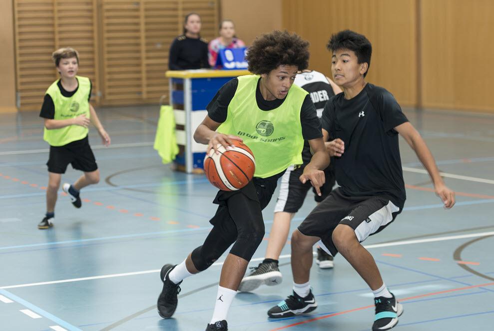 Jugendliche spielen Basketball in einer Sporthalle.