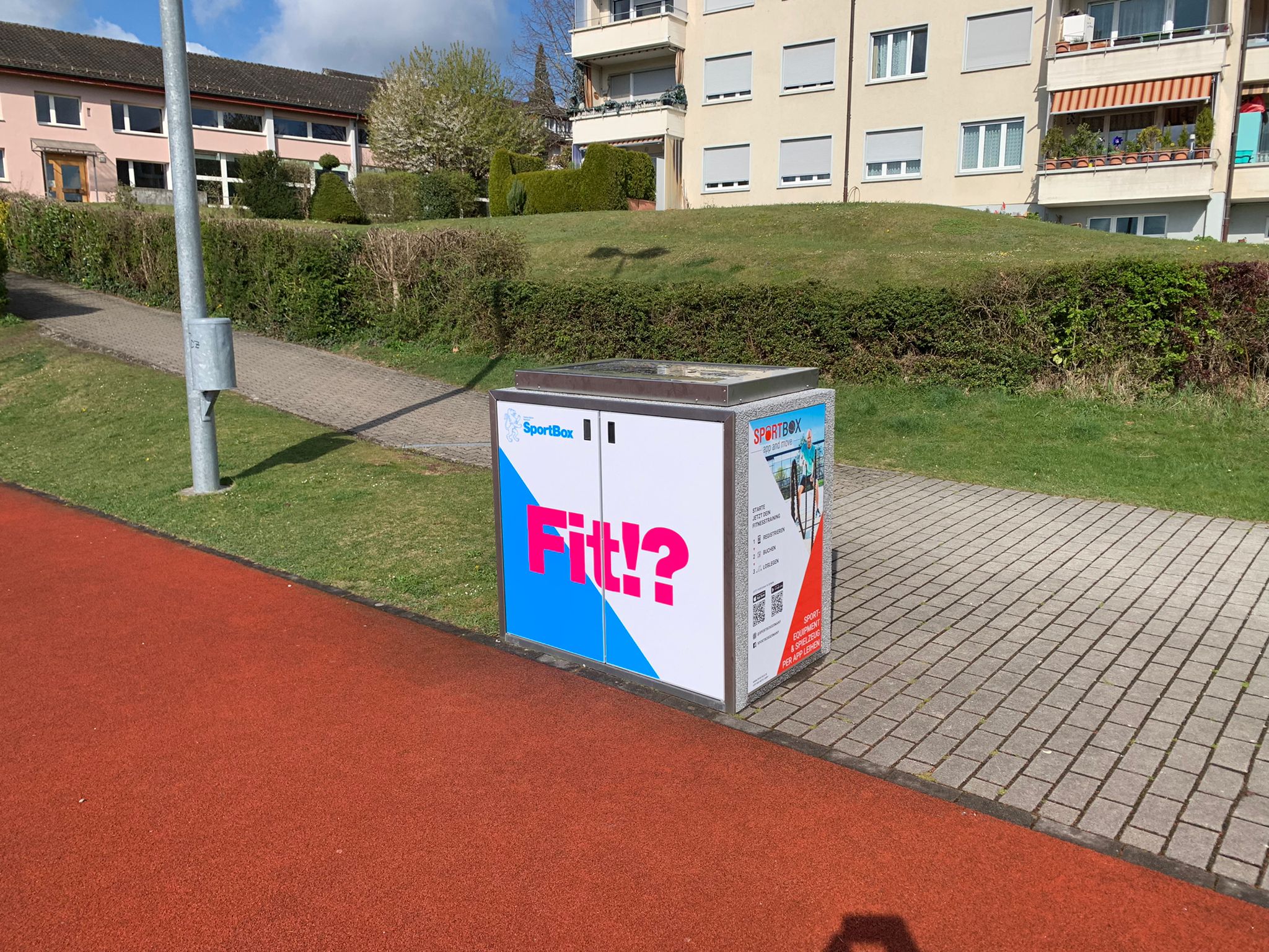 Eine SportBox (Verleihsystem mit Sport- und Fitnessequipment) steht bei einem Hartplatz in Egg, im Hintergrund sind Wohnhäuser sichtbar