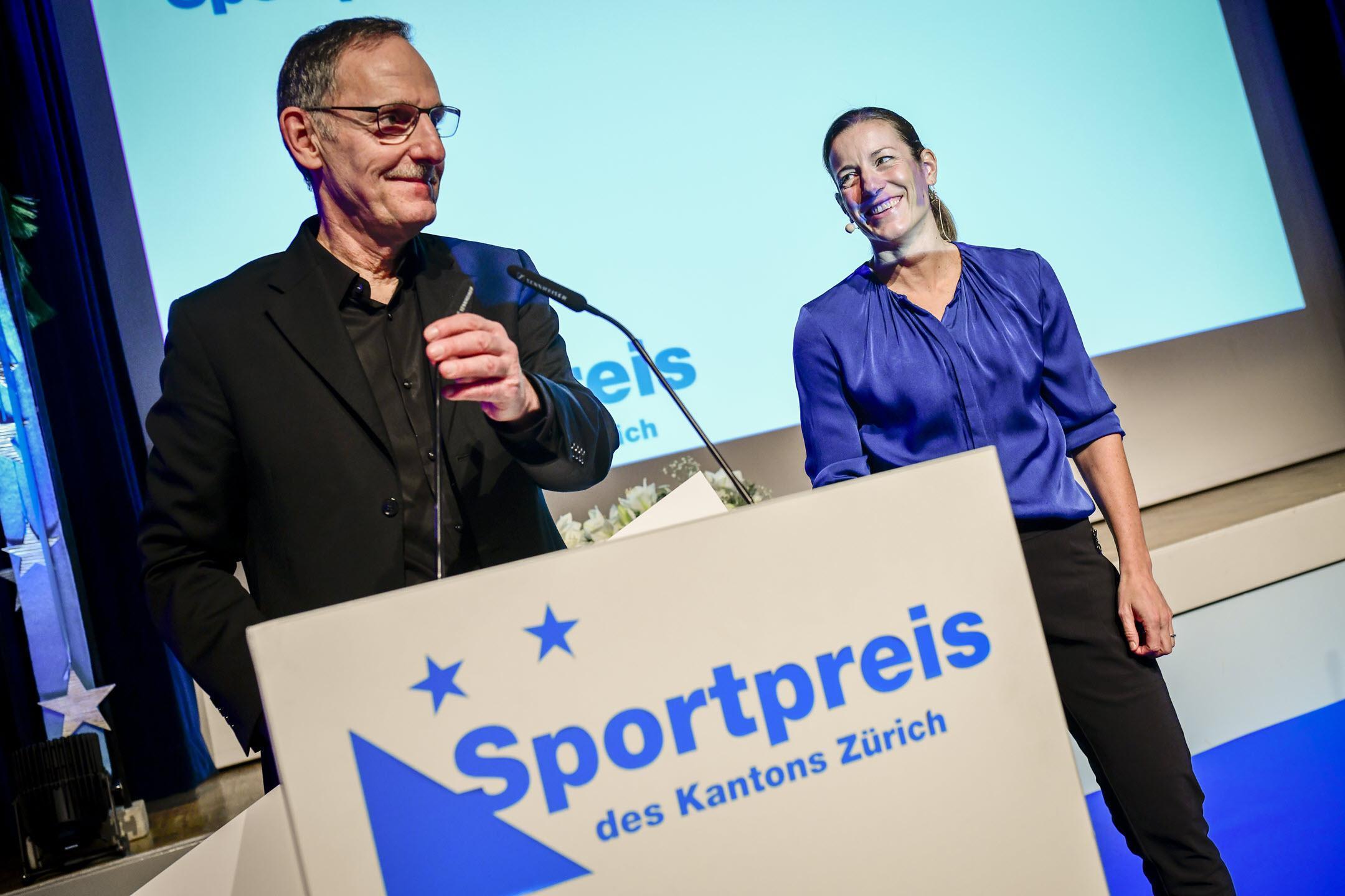 Laudatio von Regierungsrat Mario Fehr an der Verleihung des Sportpreises des Kantons Zürich 2022: Regierungsrat Mario Fehr spricht am Rednerpult, Nicola Spirig steht mit lachendem Gesicht im Hintergrund 