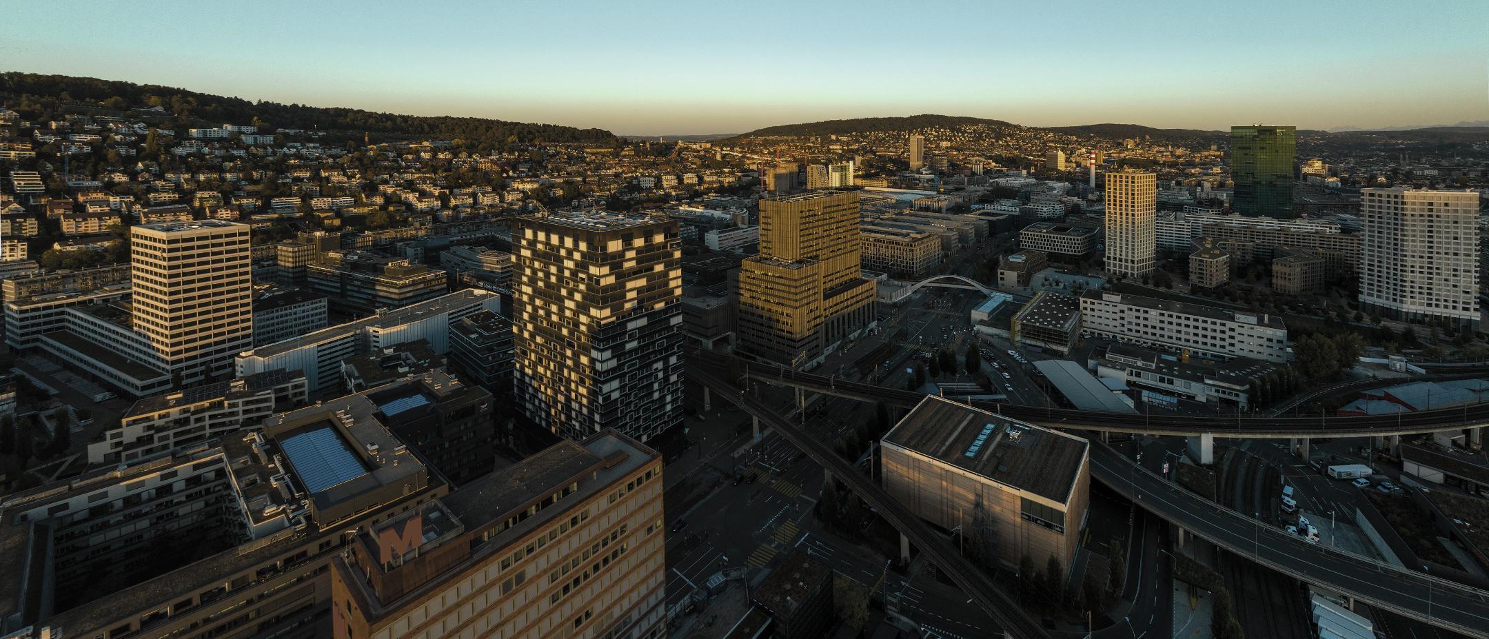 Skyline von Zürich bei Sonnenuntergang von oben, mit Gebäuden, Hügeln und weitem Himmel.