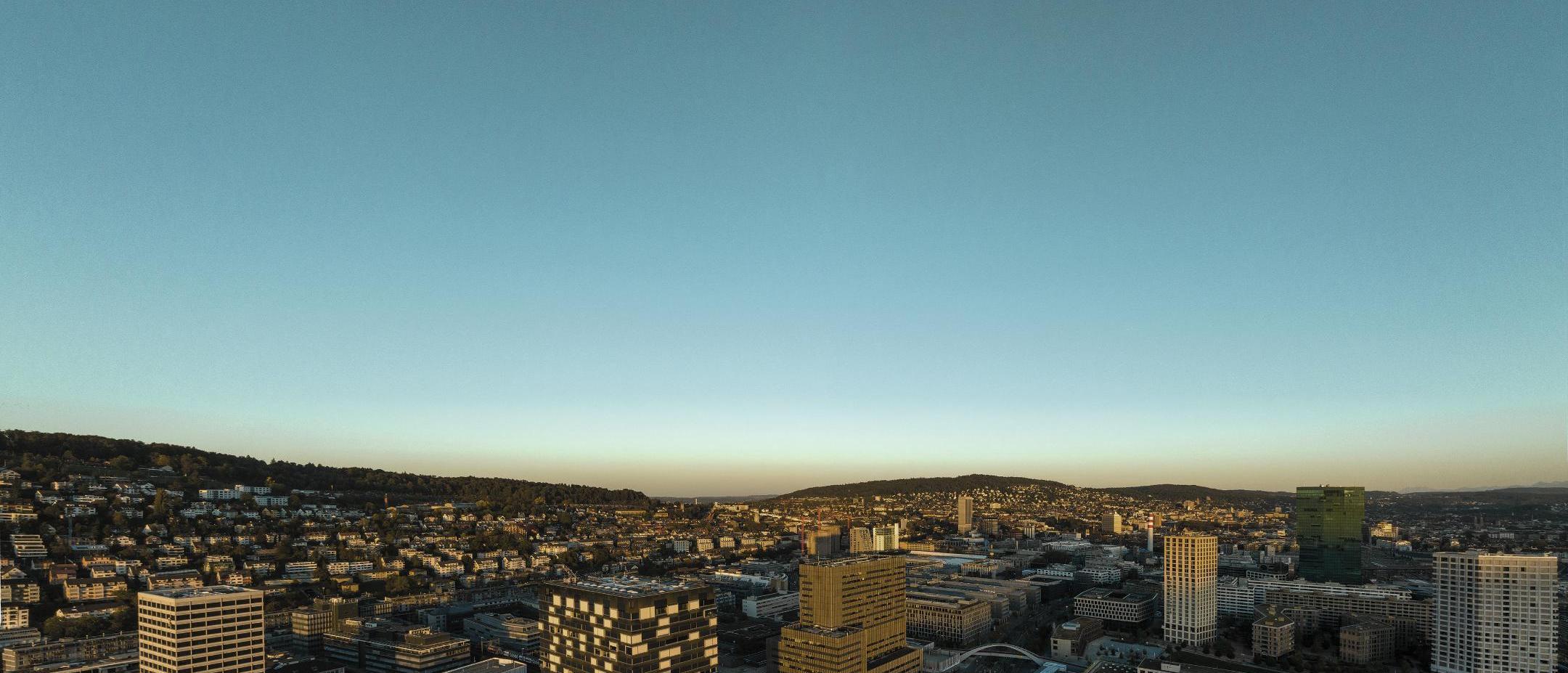 Skyline von Zürich bei Sonnenuntergang von oben, mit Gebäuden, Hügeln und weitem Himmel.