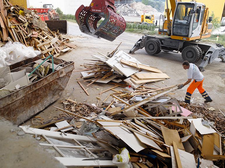 Bausperrgutsortieranlage: Holz und weitere Abfälle auf einzelnen Haufen separiert,  ein Mitarbeiter in orangen Leuchthosen hilft von Hand zu sortieren