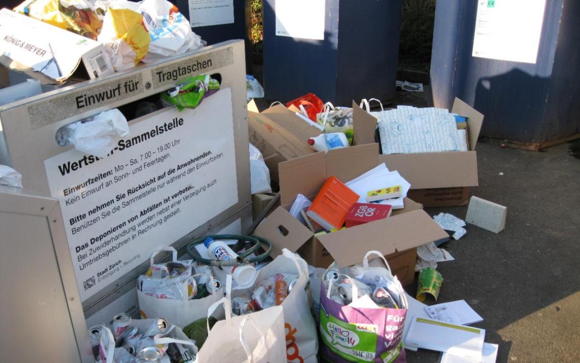 Bild einer illegalen Ablagerung von Abfällen an einer öffentlichen Sammelstelle: Kartonschachteln und Papiersäcke wurden neben den Abfallcontainern stehengelassen.